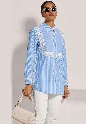 Woman wearing blue and white striped long-sleeve blouse with lace details, white pants, sunglasses, and holding a beige handbag.