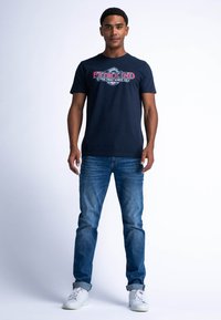 Námořnické tričko s červeným a bílým potiskem "PETROLIND". V kombinaci se světle modrými džíny a bílými teniskami, stojící na světlém pozadí.