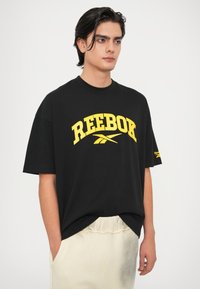Melnas kokvilnas T-krekls ar izteiksmīgu dzeltenu "Reebok" logo, brīvs piegriezums, apaļa apkakle un īsas piedurknes. Aksentēts ar logo detaļu uz piedurknes.