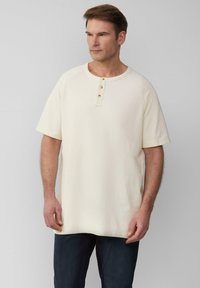 Jasno beżowa, krótka koszulka henley z krótkim rękawem, wykonana z teksturowanej tkaniny, z okrągłym dekoltem i trzema złotymi guzikami wzdłuż góry.