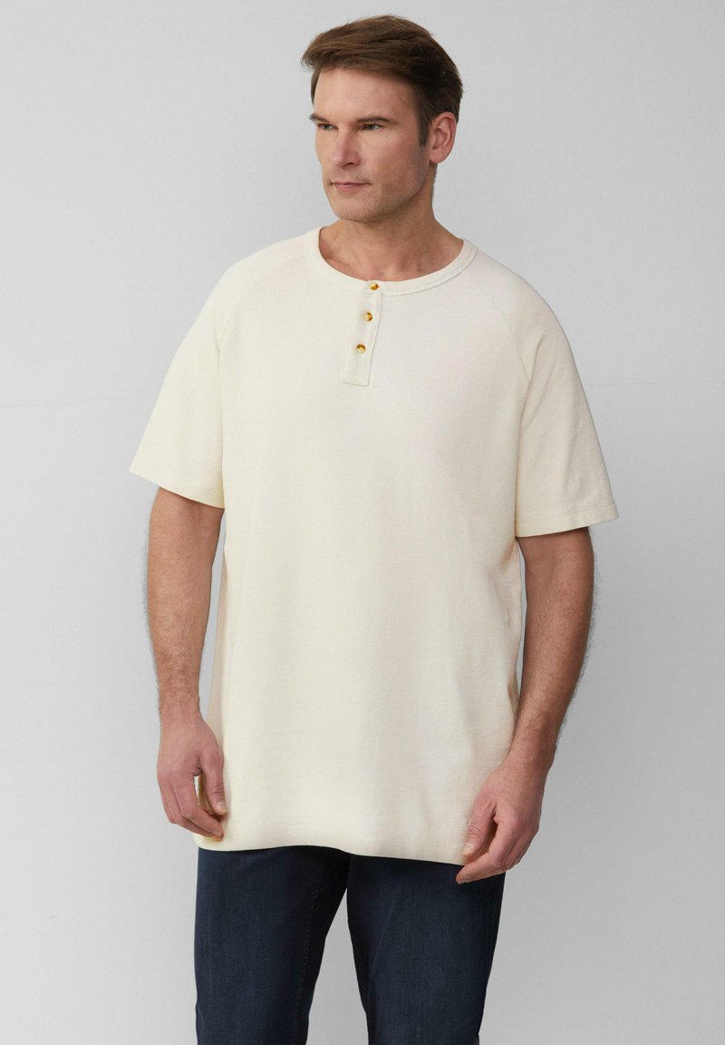 Jasno beżowa, krótka koszulka henley z krótkim rękawem, wykonana z teksturowanej tkaniny, z okrągłym dekoltem i trzema złotymi guzikami wzdłuż góry.