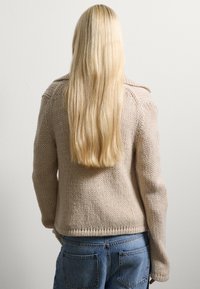 Beige knitted sweater with a textured pattern, ribbed hem, and long sleeves, paired with blue high-waisted jeans. Hair flows down the back.