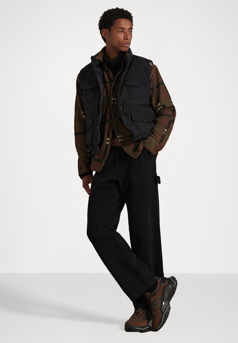 Gilet imbottito nero sopra una camicia a quadri marrone, pantaloni neri larghi e scarpe da escursionismo marroni. Caratterizzato da tasche e un colletto alto.