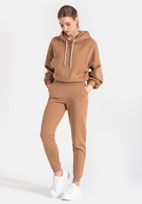 Brązowy krótki hoodie z kieszenią z przodu i białymi sznurkami, zestawiony z dopasowanymi zwężającymi się dresami i białymi sneakersami. Gładka tekstura.