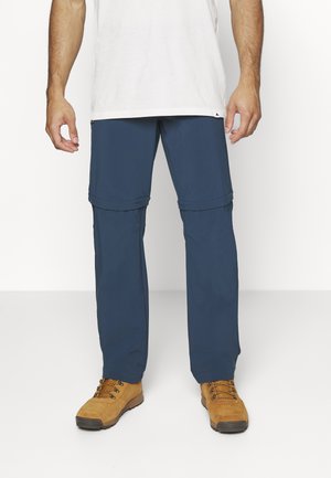 Navyblauwe cargobroek met ritsen op de knieën om om te bouwen tot shorts, met een rechte snit en lichtgewicht stof. Bruine wandelschoenen aan.