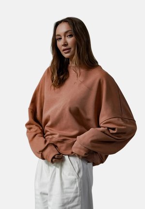 Kobieta z długimi brązowymi włosami, ubrana w luźną brązową bluzę i białe spodnie, ręce poprawiające dół bluzy.