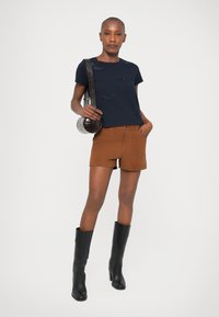 Marineblaues T-Shirt mit gemusterten Details, braune Wildledershorts und kniebhohe schwarze Lederstiefel. Beinhaltet eine runde, strukturierte Handtasche.