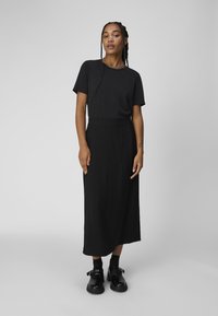 Sort T-shirt med kort ærme, parret med en sort knælang nederdel med en glat tekstur. Sorte klodsede sko fuldender outfittet.