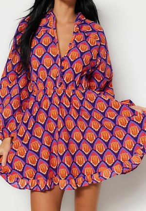 Kvinde iført en lilla kjole med orange blomsterprint, lange ærmer, knapper på brystet og en flæset kant, der holder skørtet ud.