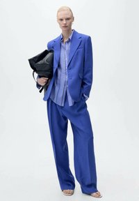 Blazer blu su misura e pantaloni a gamba ampia abbinati a una camicia a righe; tiene una borsa tote nera. Tessuto liscio, design strutturato.