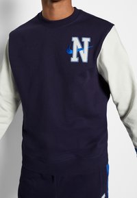 Navy sweatshirt with white sleeves, featuring a blue embroidered "N" logo and Nike swoosh on the chest. Soft fabric with a crew neck design.