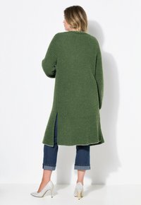 Lång, grön stickad cardigan med sidoslitsar, texturerad tyg, rund halsringning, bärs med uppvikta blå jeans och silverfärgade högklackade skor.