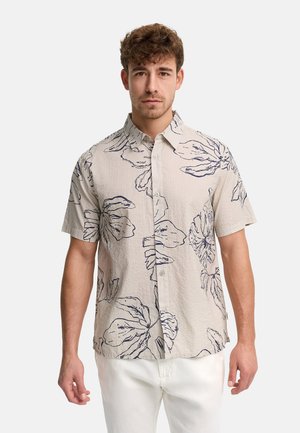 Junger Mann trägt ein beiges kurzärmliges Hemd mit großen dunkelblauen Blumenmustern und weiße Hose, steht vor einem schlichten Hintergrund.