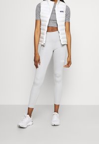 Vit pufferväst, grå t-shirt med kort ärm och ljusgrå leggings; funktioner inkluderar en figursydd design och en logotyp på benet. Vita sneakers fulländar outfiten.