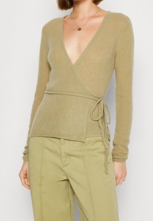 Femme portant un pull léger marron clair noué à la taille et un pantalon beige taille haute, avec un collier pendentif rond visible.