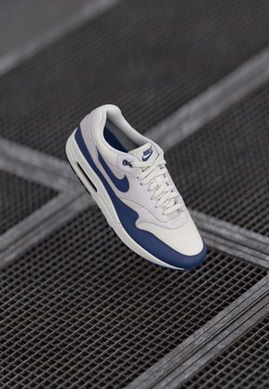 Baskets Nike Air Max blanches et bleues avec lacets, flottant au-dessus d'un sol métallique grillagé avec des lignes qui se croisent.