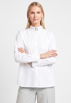 Donna bionda con orecchini a cerchio d'argento che indossa una camicia bianca abbottonata con colletto con fibbia e pantaloni grigio chiaro, braccia incrociate.