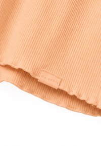 Tissu côtelé orange clair avec une petite étiquette rectangulaire cousue près de l'ourlet ondulé, portant la mention "OH APRIL" en majuscules.