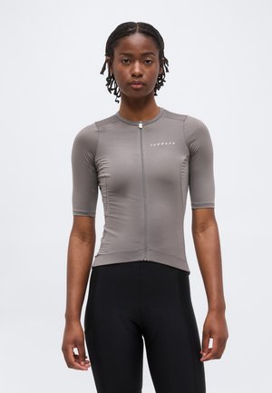 Fonott hajú nő, aki szűk, szürke, rövid ujjú kerékpáros mezben áll, elöl cipzárral és fekete kerékpáros nadrágot visel, fehér háttér előtt.