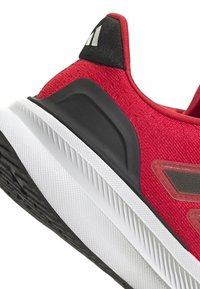 Zapatilla deportiva roja con parte superior texturizada, acentos negros y una suela blanca con bordes estriados y un fondo con patrón.