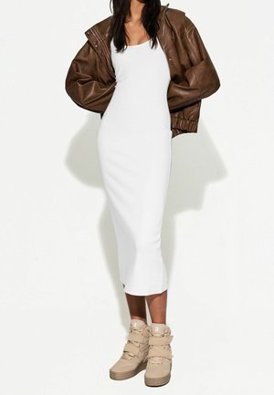 Kobieta w białej dopasowanej sukience midi bez rękawów, oversize'owej brązowej skórzanej kurtce i beżowych wysokich trampkach, stojąca na białym tle.