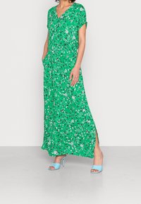 Nainen, joka on pukeutunut vihreään kukkakuosiseseen maksimekkoon, jossa on taskut, sekä vaaleansinisiin korkokenkiin, seisoo harmaalla lattialla.