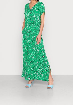 Nainen, joka on pukeutunut vihreään kukkakuosiseseen maksimekkoon, jossa on taskut, sekä vaaleansinisiin korkokenkiin, seisoo harmaalla lattialla.