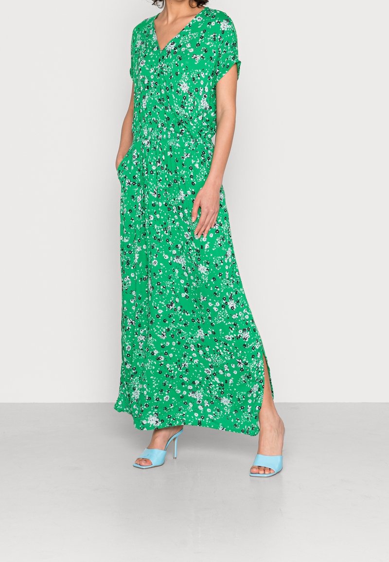 Nainen, joka on pukeutunut vihreään kukkakuosiseseen maksimekkoon, jossa on taskut, sekä vaaleansinisiin korkokenkiin, seisoo harmaalla lattialla.