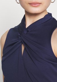 Femme portant un haut sans manches bleu marine avec un détail de nœud torsadé au niveau de l'encolure et des boucles d'oreilles créoles dorées.