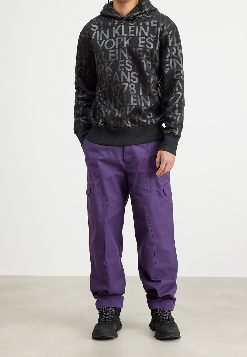 Black hoodie with reflective logo text, paired with loose purple cargo pants featuring side pockets. Combined with black sneakers.