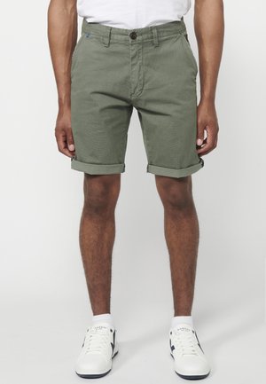 Shorts di cotone verdi con orli arrotolati, dotati di chiusura anteriore con bottone e tasche laterali, abbinati a sneakers bianche.