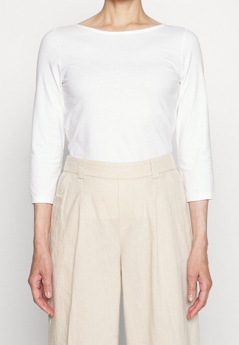 Person som bär en vit långärmad topp instoppad i högmidjade, beige vida byxor med bälteshällor och sidofickor.