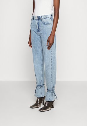 Pantalones de denim azul claro con un corte relajado, que cuentan con puños anudados, bolsillos frontales y un cierre de botón. Llevados con botas de tacón y punta.