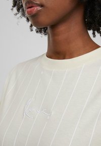 Camiseta de color crema con rayas verticales blancas, cuello redondo y sutil logo bordado. Tejido suave con textura lisa.