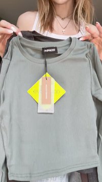 Camisola de manga longa de cor verde-claro com detalhes em frisos, com um decote redondo, etiqueta pendurada, segurada entre um item cinza e a camisa de uma pessoa.