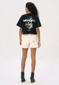 Zwart cropped t-shirt met witte tekst en grafisch ontwerp op de achterkant, gecombineerd met beige korte broek met achterzakken. Model draagt zwarte hoge hakken.