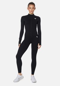 Schwarzes Sportoberteil mit hohem Kragen und durchgehendem Reißverschluss, kombiniert mit schwarzen Leggings. Beide haben eine taillierte Passform und ein Logo auf der Brust.