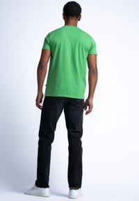 Camiseta de algodón verde con cuello redondo, combinada con jeans negros. Persona lleva zapatillas blancas. Vista desde atrás contra un fondo claro.