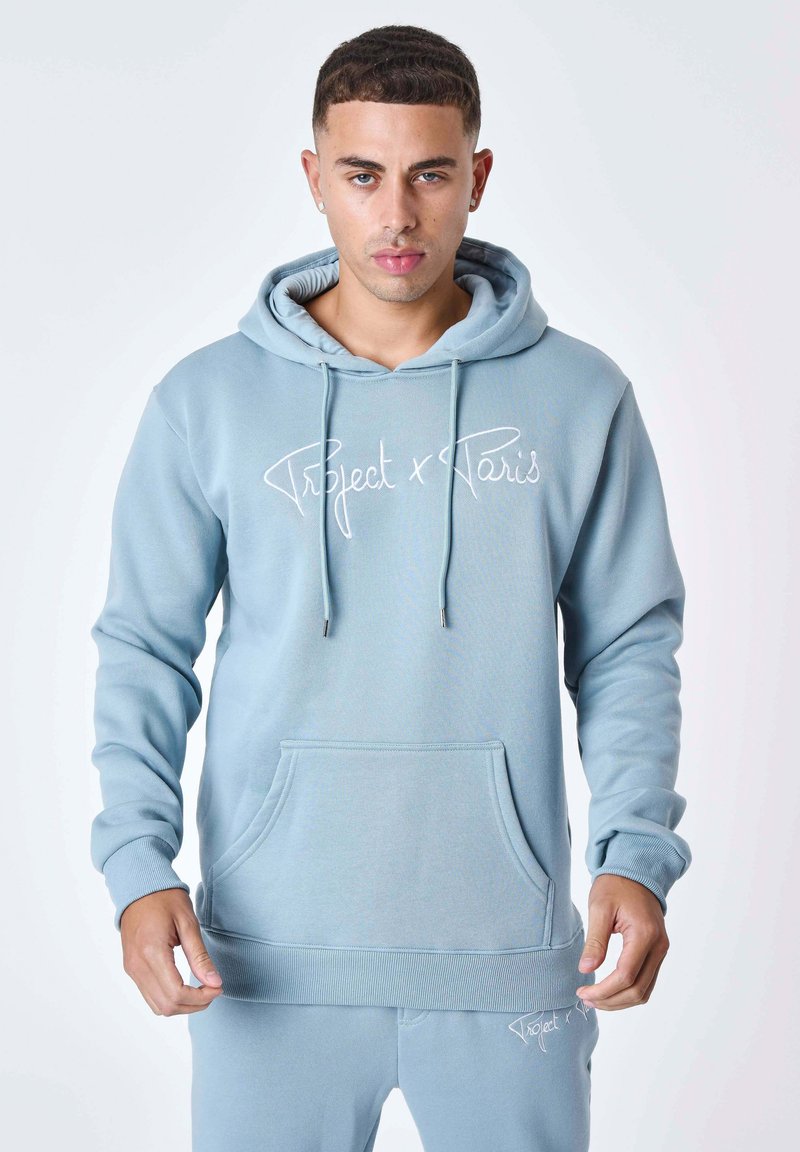 Jeune homme aux cheveux courts portant un sweat à capuche bleu clair et un pantalon assorti, debout devant un fond blanc uni.