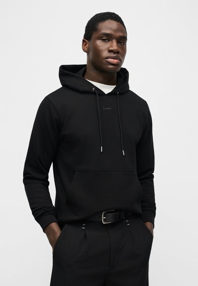 Νέος άνδρας που φοράει μαύρο φούτερ με κουκούλα και τσέπη μπροστά και μαύρο παντελόνι, όρθιος με τα χέρια στις τσέπες μπροστά σε απλό φόντο.