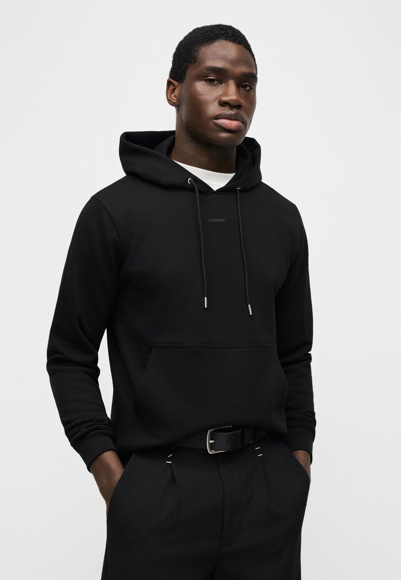 Junger Mann trägt einen schwarzen Kapuzenpullover mit Fronttasche und schwarze Hose, steht mit den Händen in den Taschen vor einem einfarbigen Hintergrund.