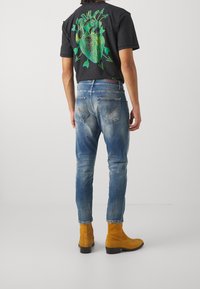 Svart t-shirt med en grön grafisk design, kombinerad med ljusblå denimjeans. Outfitten kompletteras med gula mockastövlar.