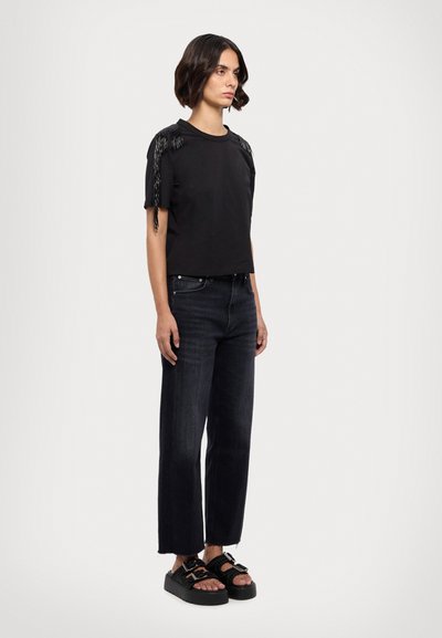 Chemise noire à manches courtes avec des franges sur les épaules, assortie à un pantalon en denim large de couleur foncée et des sandales noires à semelles épaisses.