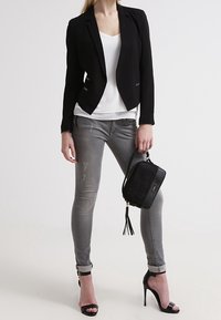 Černý sak na míru přes bílé tričko, spárovaný se šedými slim-fit džíny s mírným opotřebením a černými sandály na vysokém podpatku. Černá kabelka s třásněmi.