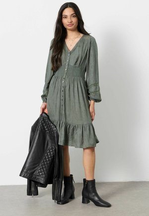 Woman with long dark hair wears a green buttoned dress and black ankle boots, holding a black quilted leather jacket in one hand.