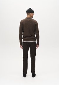 Bruine wollen trui met een ronde hals, gecombineerd met donkere, taps toelopende broek en zwarte schoenen, gezien van achteren. Zwarte beanie op het hoofd.