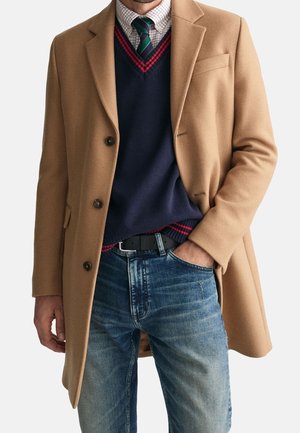 Mann trägt einen Kamelhaarmantel über einem marineblauen Pullover mit rotem Besatz, ein kariertes Hemd, grün gestreifte Krawatte und blaue Jeans mit der Hand in der Tasche.