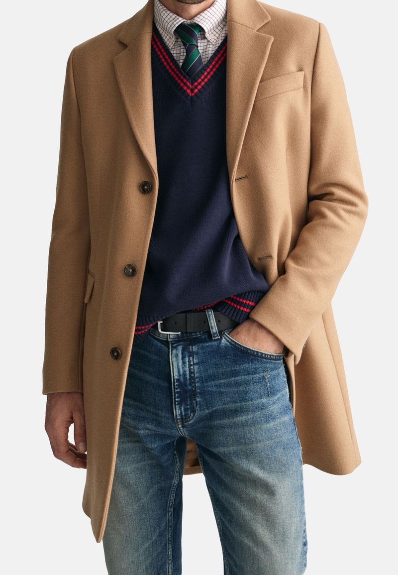 Homme portant un manteau camel par-dessus un pull bleu marine à bordure rouge, une chemise à carreaux, une cravate rayée verte, et un jean bleu avec la main dans la poche.