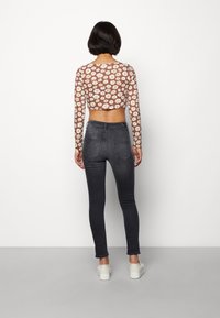 Mujer de espaldas vestida con una blusa corta de manga larga con estampado de margaritas, jeans skinnies oscuros y zapatillas blancas, con un fondo blanco liso.