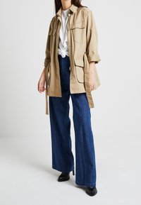Beige utilityjack met vier zakken aan de voorkant, opgerolde mouwen en een strik in de taille, gecombineerd met een wijd gesneden donkerblauwe jeans en zwarte enkellaarsjes.